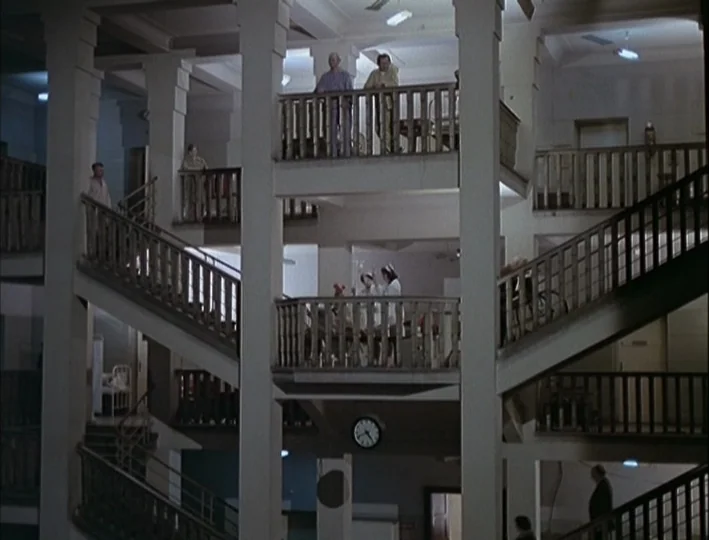 Camera Buff (1979) movie still: wide — A multi-story hospital atrium features concrete pillars, wooden balustrades, and connecting staircases.…