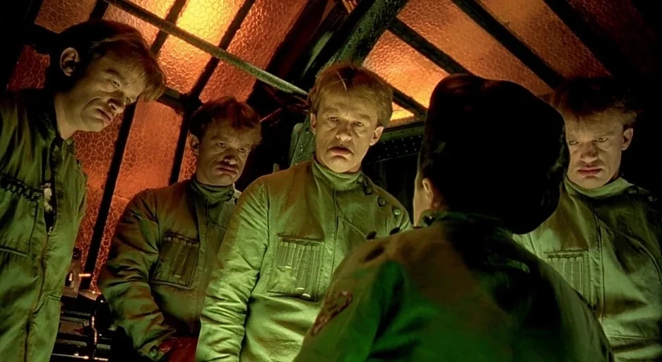 The City Of Lost Children movie still: medium — A dark, cramped metallic room features glowing amber panels overhead and complex structural beams.…