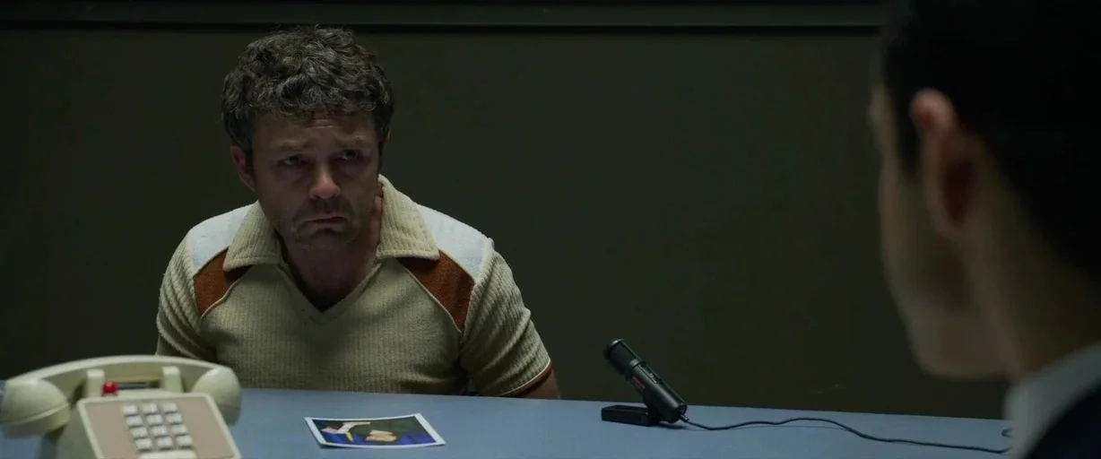 The Little Things (2021) movie still: medium — A dimly lit room with plain walls and a metallic blue surface. A man sits at the table looking at an…