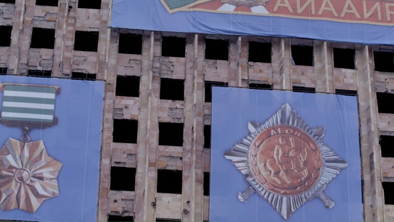 Letters to Max (2014) movie still: establishing — The exterior stone wall of a multistory building features rows of dark empty windows. Large blue…