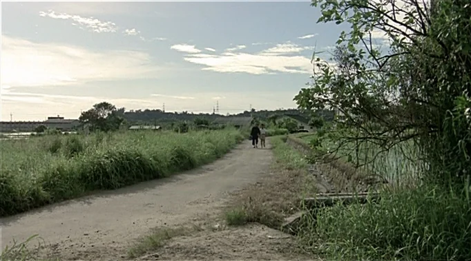 A Time To Love and A Time To Die (1985) movie still: wide — A long dirt path winds through green fields under an expansive, cloudy sky. Two people…