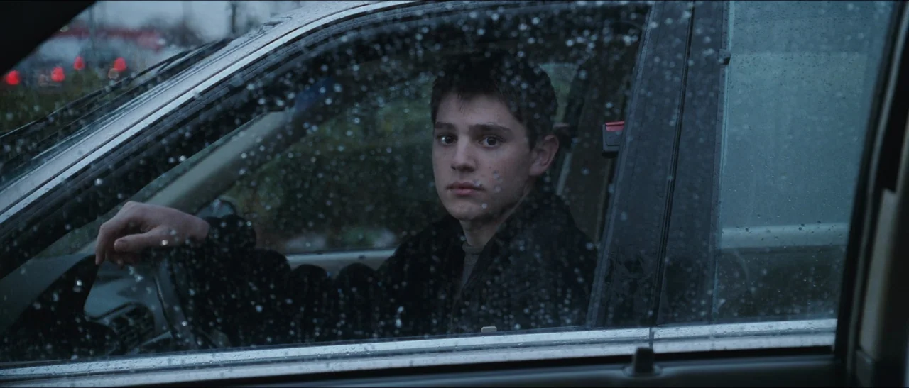 Election (1999) movie still: medium — A teenage boy sits behind the steering wheel of a car during a rainstorm. Rain droplets cover the side window…