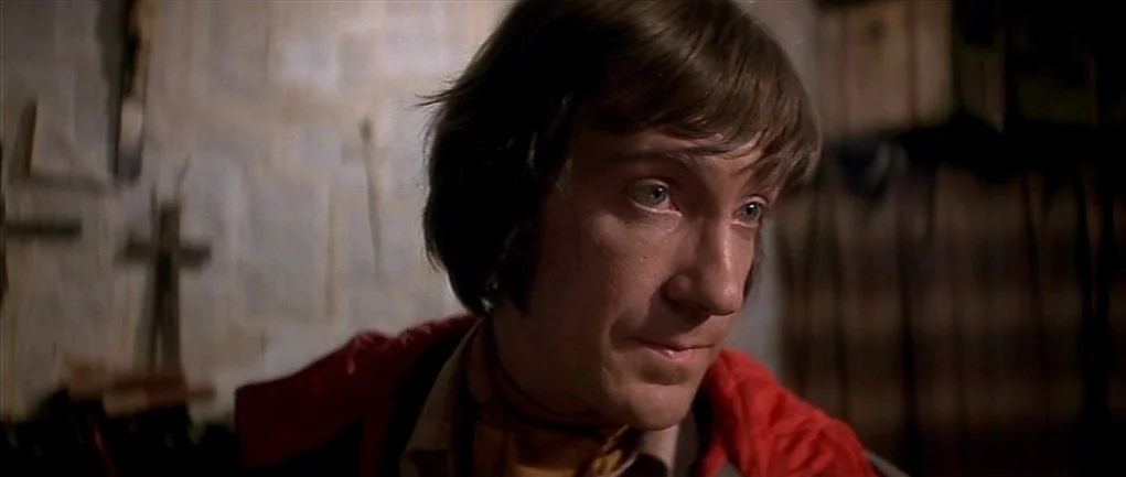 The Omen (1976) movie still: closeup — A dark workshop space features wooden tools hung against a textured wall. A man with brown hair stands inside…