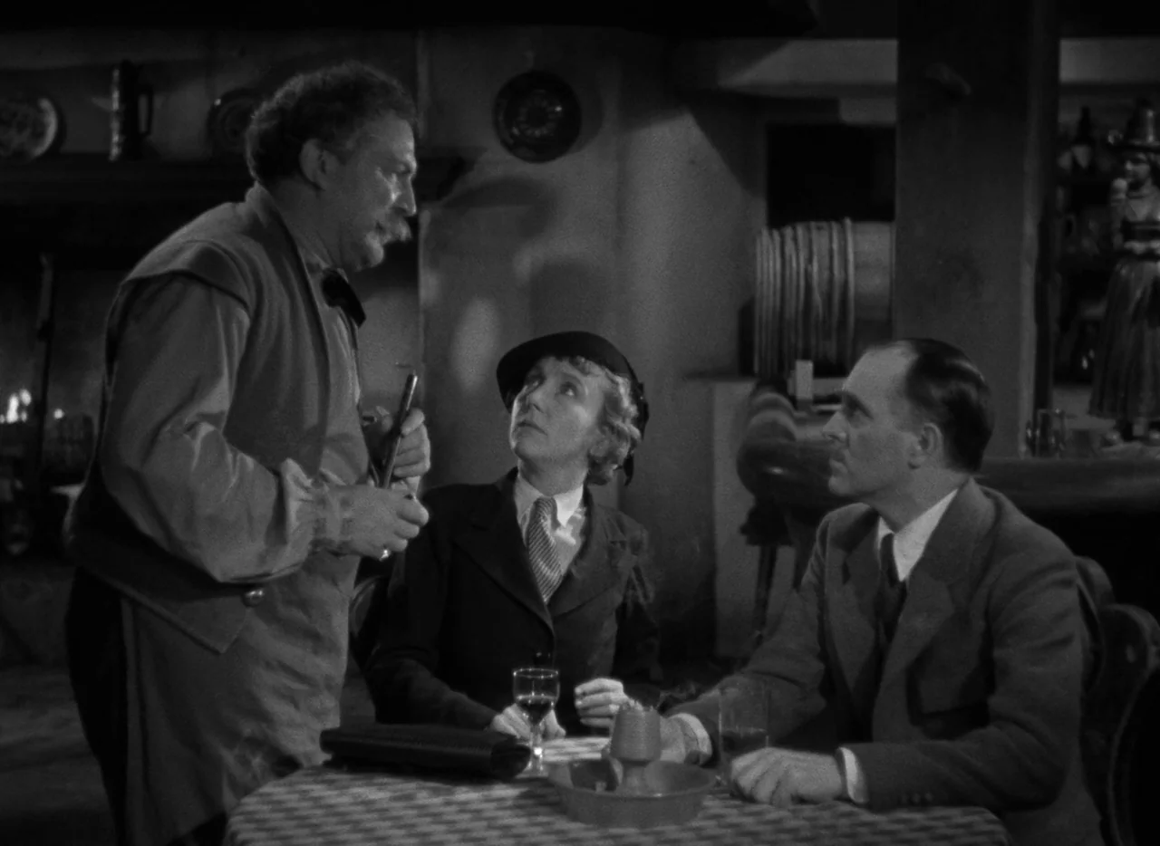 Mark of the Vampire (1935) movie still: medium — A dimly lit tavern features a wooden barrel and a fireplace in the background. A standing man talks…