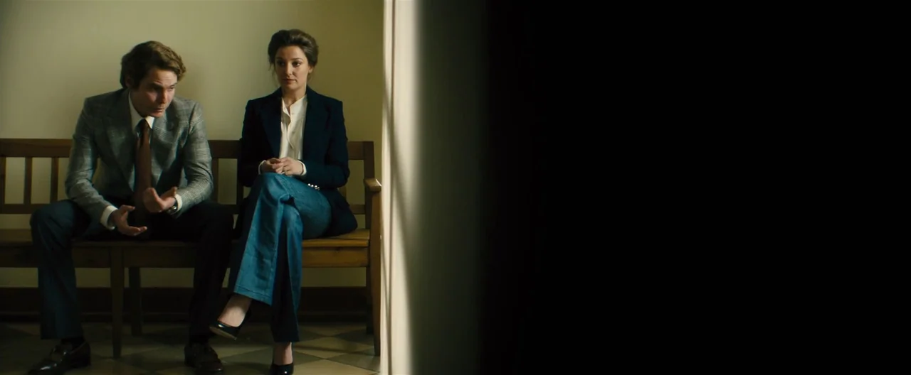 Rush (2013) movie still: medium — A man and woman sit on a simple wooden bench in a beige room. The man gestures with his hands while speaking to…