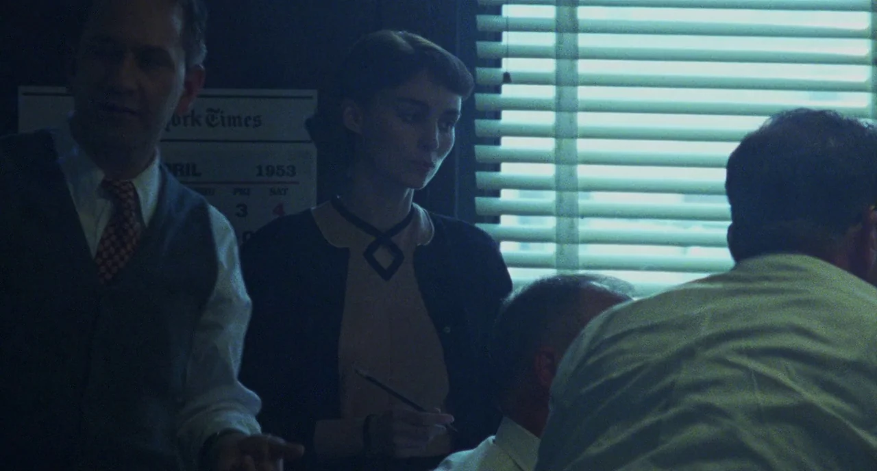 Carol (2015) movie still: medium — A 1950s office space features a New York Times wall calendar from April 1953. A woman stands between two seated…