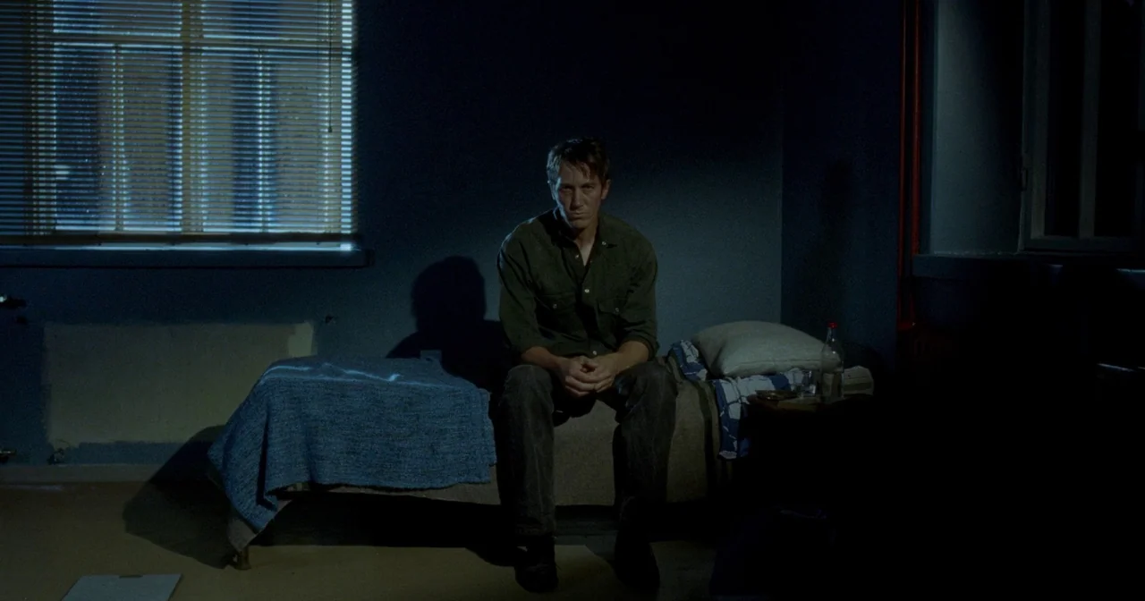 Fallen Leaves (2023) movie still: medium — A sparsely furnished room with dark blue walls features a single bed by a window. A man in a dark green…