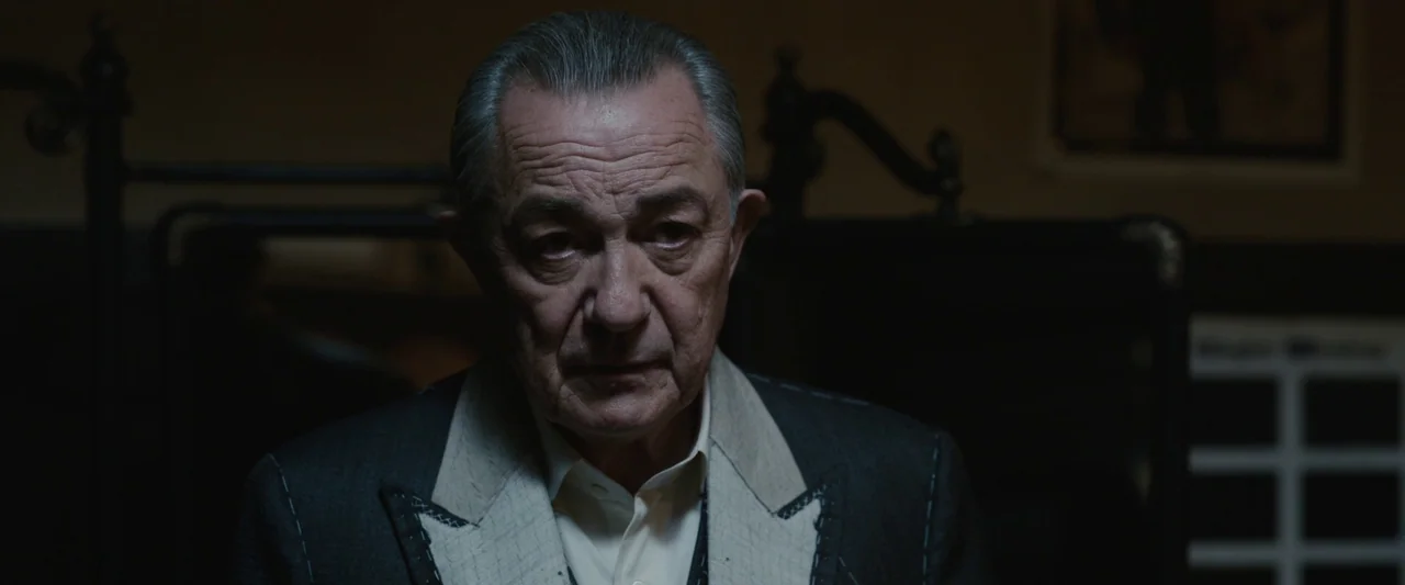 Live By Night (2016) movie still: closeup — A dark space features a man in a bespoke tailored suit jacket. He stands facing the camera with a stoic…