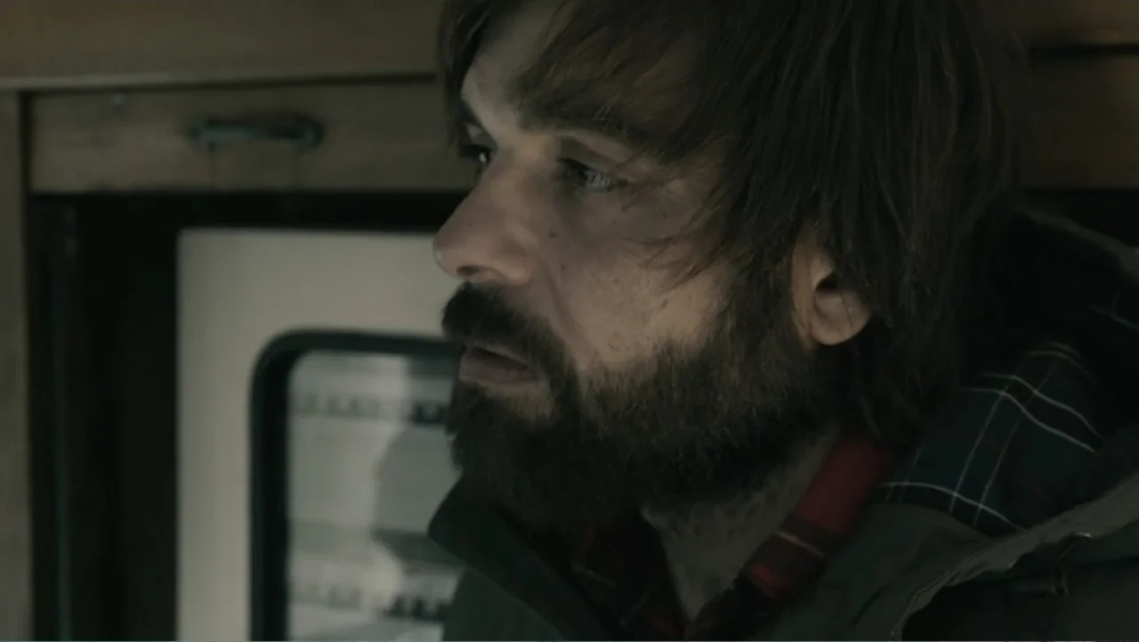 Hondar 2050 (2018) movie still: closeup — An interior space features wood paneling and a small window frame in the background. A bearded man with…