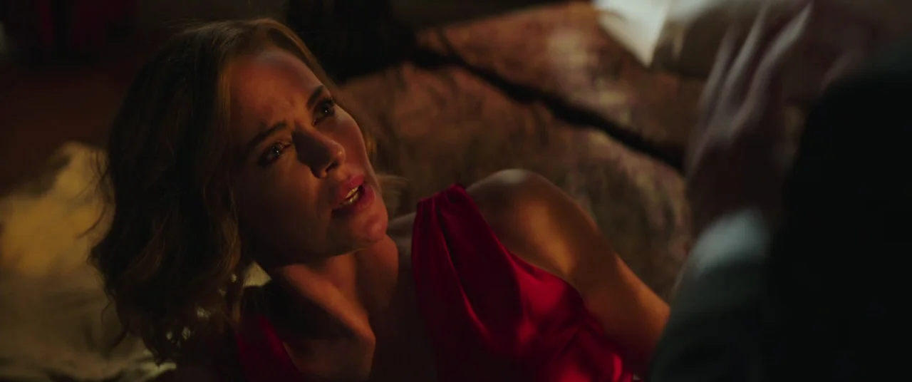 Long Shot (2019) movie still: closeup — A dimly lit hotel room features a textured headboard and muted brown bedding. A woman in a red dress…