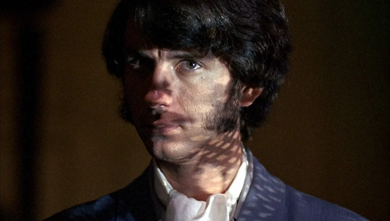 Head (1968) movie still: closeup — A dimly lit space with a dark background surrounds the subject. The man stares forward with geometric patterns of…