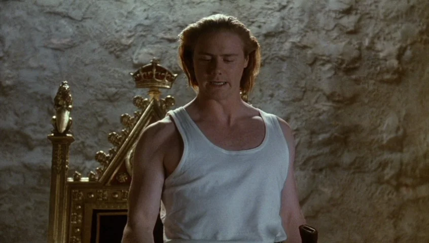 Edward II (1991) movie still: medium — A room with a rough stone wall and an ornate, high-backed golden chair. A man in a white tank top stands in…