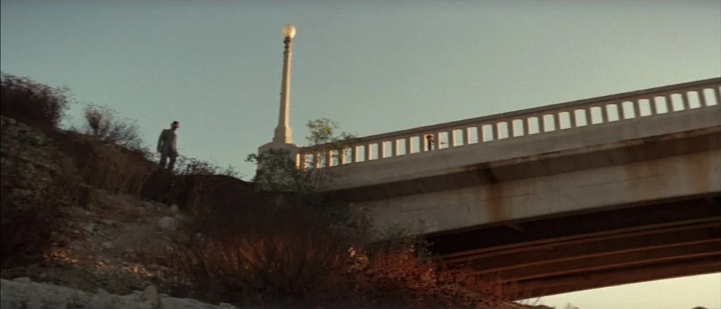 Chinatown (1974) movie still: wide — A concrete bridge spans across a dry, brush-filled embankment under a clear sky. A man in a suit stands on the…
