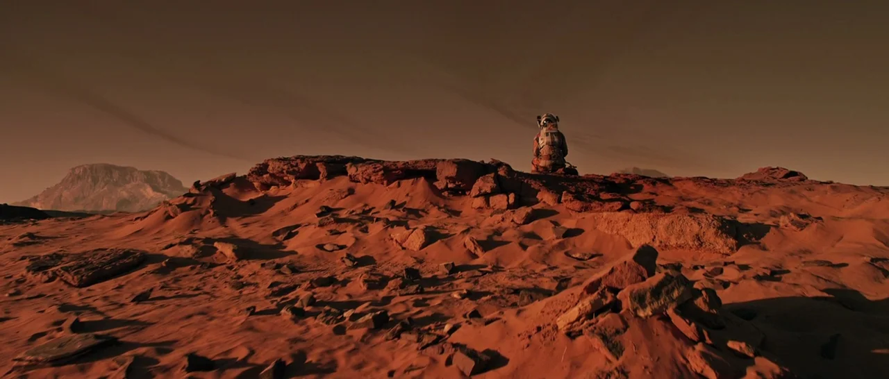 The Martian (2015) movie still: wide — A vast, arid Martian landscape features reddish sand, jagged rock formations, and distant craters. A lone…
