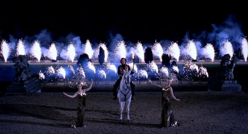 The Man Who Cried (2000) movie still: wide — A dark outdoor space is illuminated by a row of bright, sparking fireworks display. A man sits atop a…