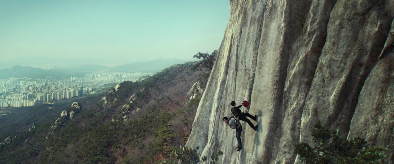Decision To Leave (2022) movie still: wide — A vertical rock cliff face rises above a forested valley overlooking a distant city. Two men in formal…