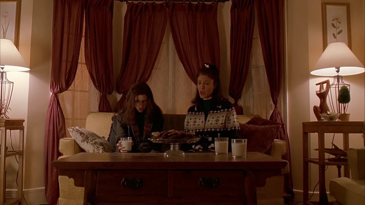 Ginger Snaps (2000) movie still: medium — A living room features a sofa with deep red curtains behind it. Two women sit on the couch with glasses of…