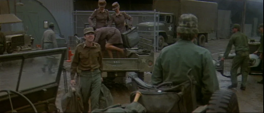 MASH (1970) movie still: medium — A military motor pool contains various green trucks and jeeps parked on dirt ground. Several soldiers in uniform…