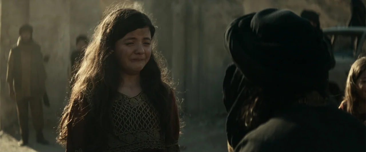 12 Strong (2018) movie still: medium — A dusty outdoor path with blurred figures standing in the background. A young girl with long dark hair cries…