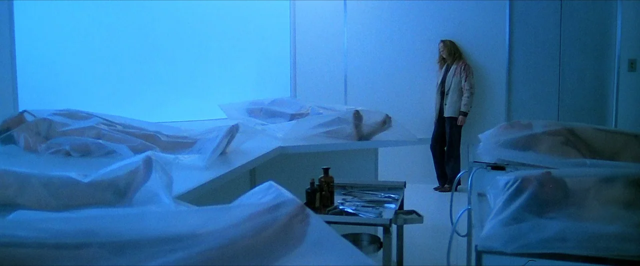 The Beyond (1981) movie still: wide — A sterile morgue features several bodies covered by thin translucent plastic sheets on tables. A woman in a…