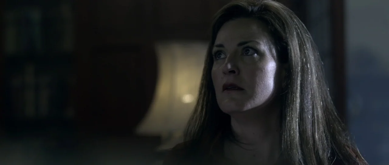 The Lullaby (2019) movie still: closeup — A dimly lit interior space features a blurred lamp in the background. A woman looks off-camera with a…