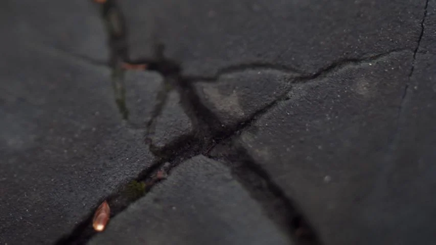 Touchy Feely (2013) movie still: closeup — A close view of a dark, weathered concrete surface with deep intersecting cracks. The cracks reveal…