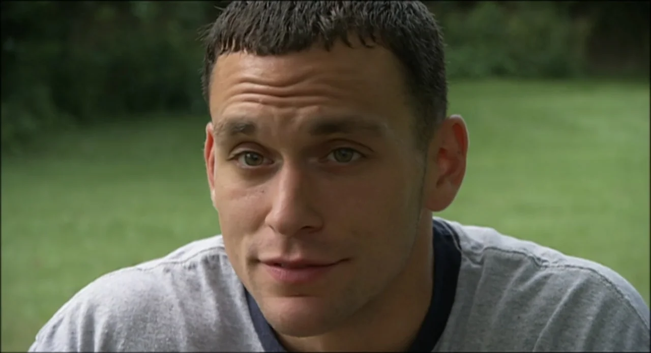 Hard as Nails (2001) movie still: closeup — An outdoor lawn setting with blurred green vegetation in the background. A young man wearing a grey…