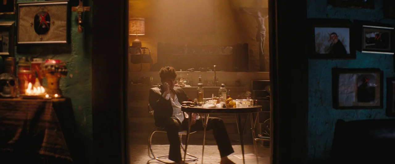 Bullet Train (2022) movie still: wide — A dimly lit room with teal walls features numerous framed photos marked with red crosses. A man sits at a…