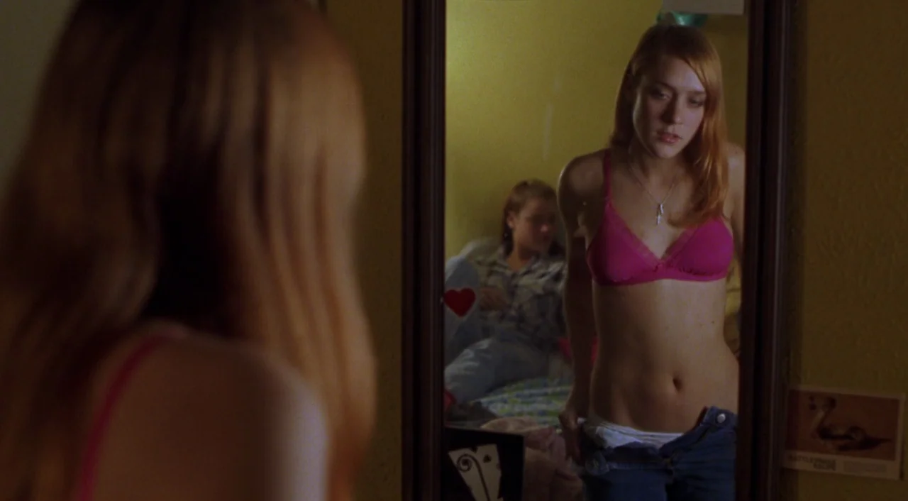 Boys Don’t Cry (1999) movie still: medium — A teenage girl stands in front of a mirror in a yellow-walled bedroom. She examines her reflection while…
