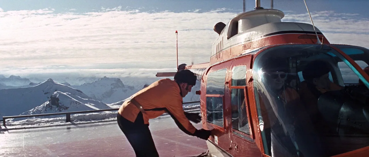 On Her Majesty’s Secret Service (1969) movie still: fullbody — A red and white helicopter sits on a frozen pad atop snowy mountains. A man in an…