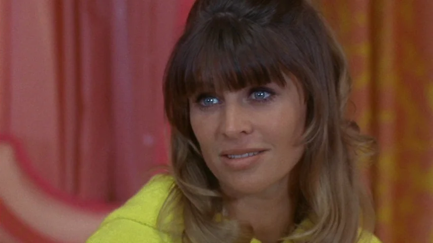 Petulia (1968) movie still: closeup — A woman stands indoors in front of textured, orange-toned curtains. She looks toward the side while wearing a…