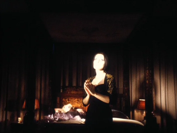 The Death of Maria Malibran (1972) movie still: medium — A dark, ornate bedroom featuring a large canopy bed with heavy dark curtains. A woman in a…