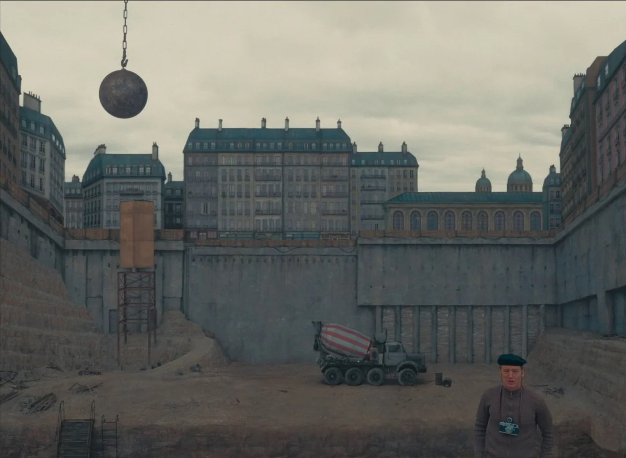 The French Dispatch (2021) movie still: wide — A wide, flat construction pit is surrounded by tall residential buildings under overcast skies. A man…
