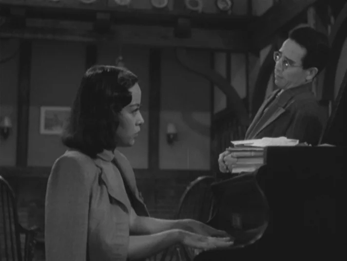 Scandal (1950) movie still: medium — An interior space features wood-paneled walls with dark timber framing and a piano. A woman sits at the piano…