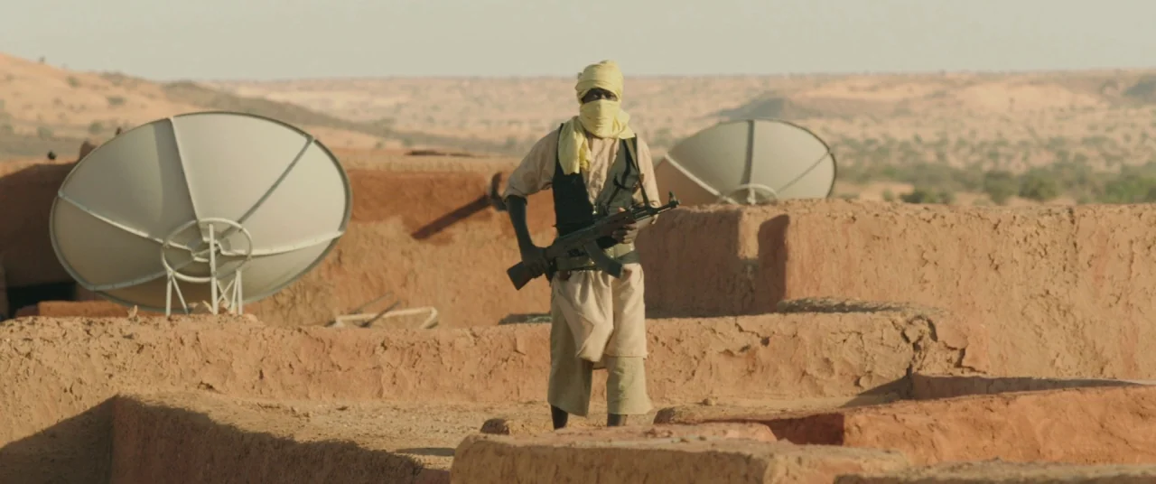 Timbuktu (2014) movie still: fullbody — A flat mud-brick rooftop overlooks a vast, arid desert landscape under a clear sky. A man stands at the…