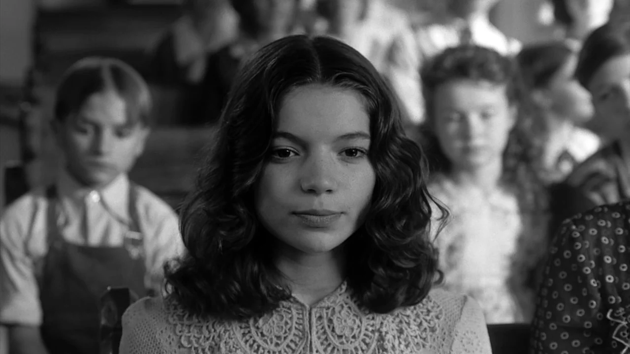 The Notorious Bettie Page (2005) movie still: closeup — A student sits at a wooden desk among other children in a school room. The girl in the…