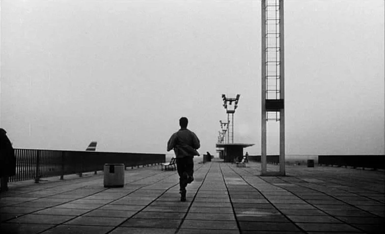 La Jetée (1962) movie still: wide — An expansive flat concrete observation deck at an airport under a grey sky. A young man runs directly away from…