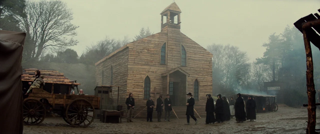 Never Grow Old (2019) movie still: wide — A rustic frontier town square features a large wooden church with a bell tower. A group of people in dark…