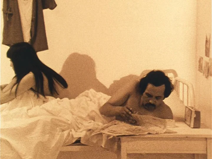 The Woman with Two Heads (1977) movie still: medium — A spare room with white walls, a single bed, and a small wooden table. A man reads a newspaper…