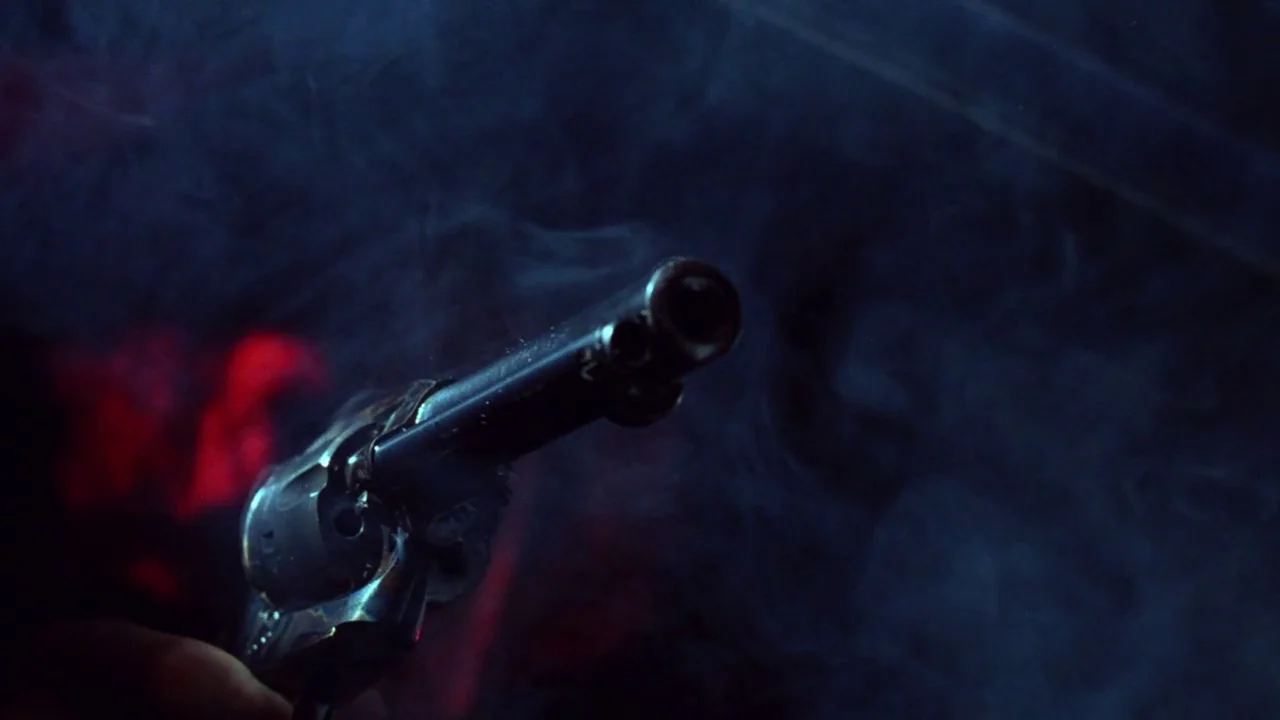The Thin Blue Line (1988) movie still: closeup — A close-up view shows the metallic barrel of a revolver against a dark background. Wisps of grey…