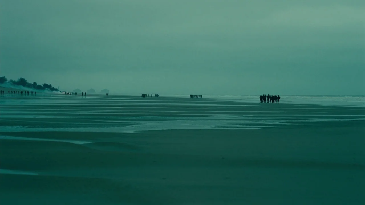 Dunkirk (2017) movie still: wide — A wide, empty expanse of wet, flat sand stretches toward an overcast sea. Several small groups of people in dark…