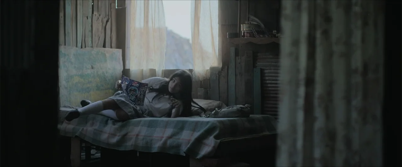 Radical (2023) movie still: medium — A small rustic room with wooden walls and a window with sheer curtains. A young girl reclines on a…