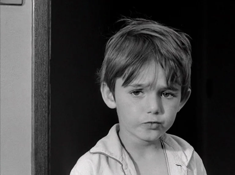 Une Femme Mariée (1964) movie still: closeup — A young boy stands in a doorway against a dark background. He faces slightly off-camera with a…