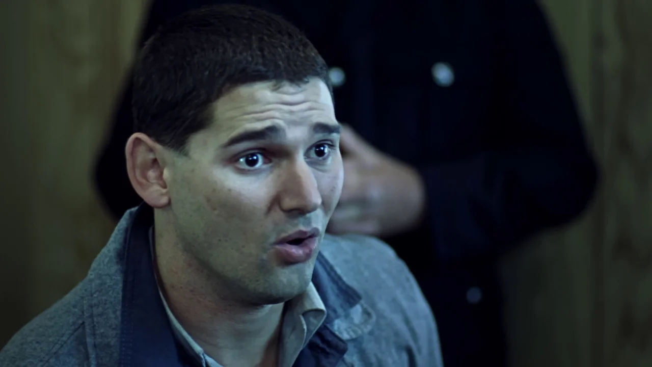 Chopper (2000) movie still: closeup — A dimly lit room features a man in the foreground speaking to an unseen person. A second person in a dark coat…