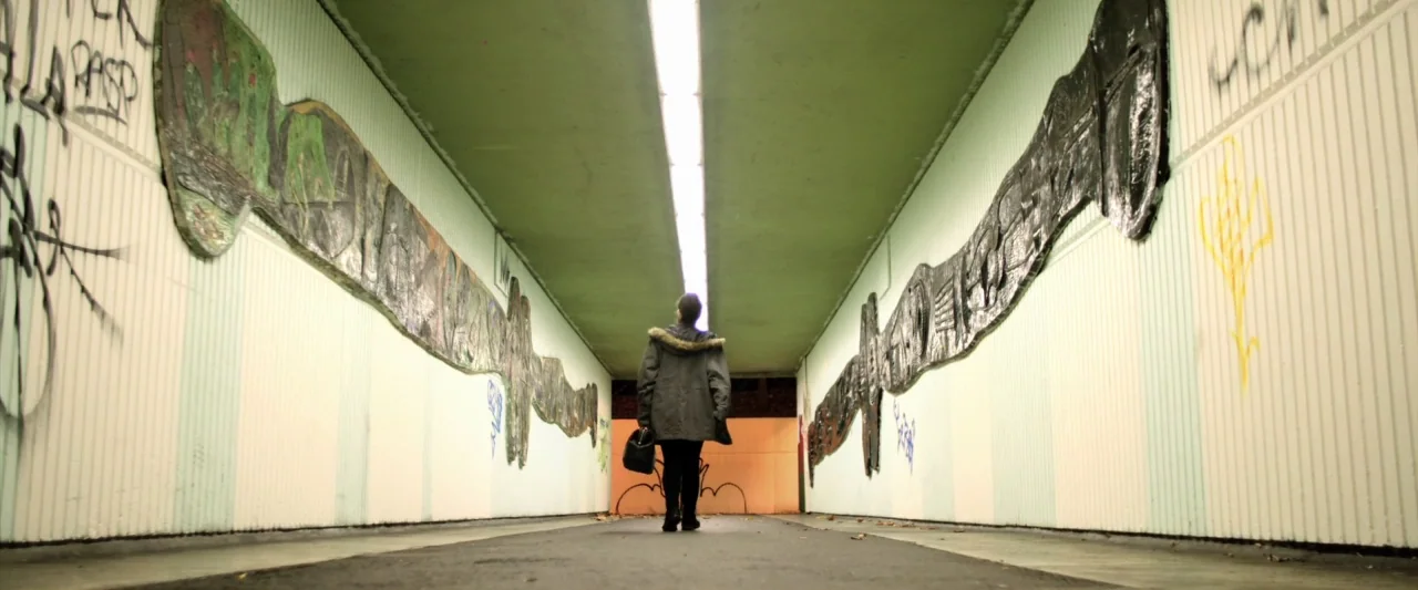 Prevenge (2016) movie still: wide — A long concrete pedestrian tunnel features white walls covered in dark raised artistic murals. A lone figure…