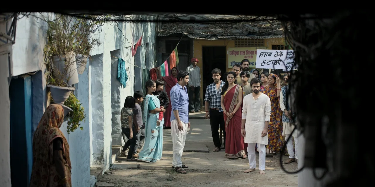 Dry Day (2023) movie still: wide — A group of people stands in a narrow village path lined with light blue walls. Several individuals are gathered…