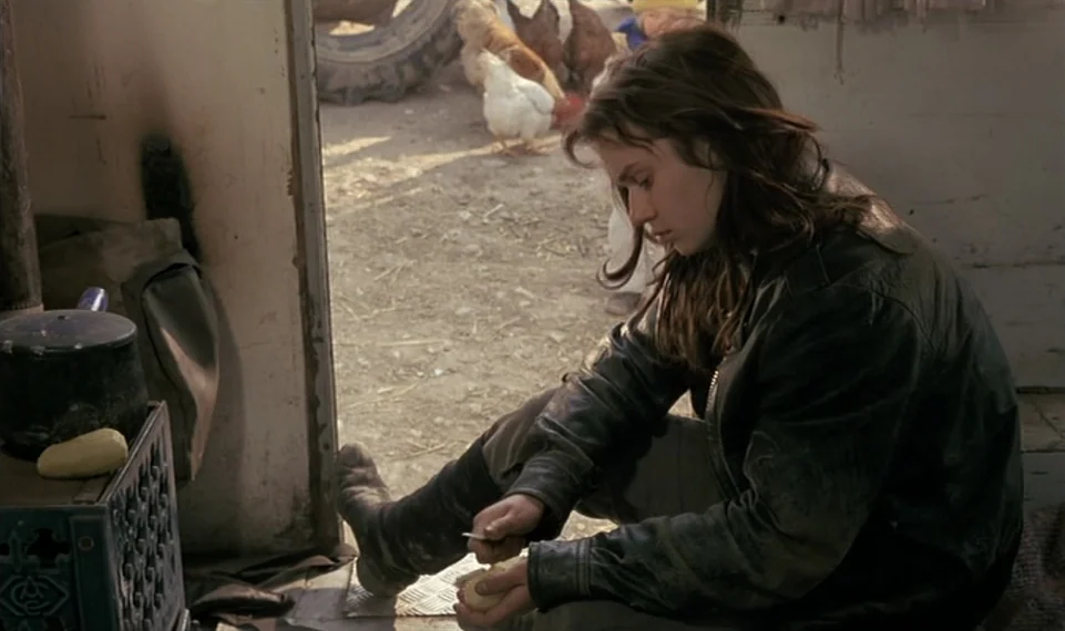 Vagabond (1985) movie still: medium — A rustic structure doorway opens onto a dirt farmyard with chickens near a tire. A young woman in a leather…
