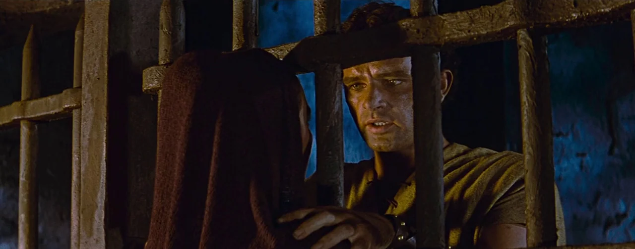The Robe (1953) movie still: medium — A dark stone cell features heavy metal bars separating the interior from the outside. A man in a simple tunic…