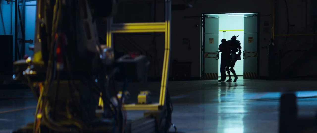 Chappie (2015) movie still: wide — A dark industrial space features an open doorway leading into a brightly illuminated room. An anthropomorphic…