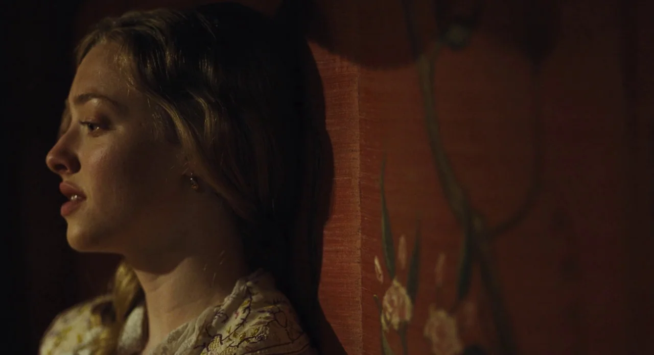 Les Misérables (2012) movie still: closeup — A dimly lit interior with a red textured wall featuring painted floral designs. A woman stands against…